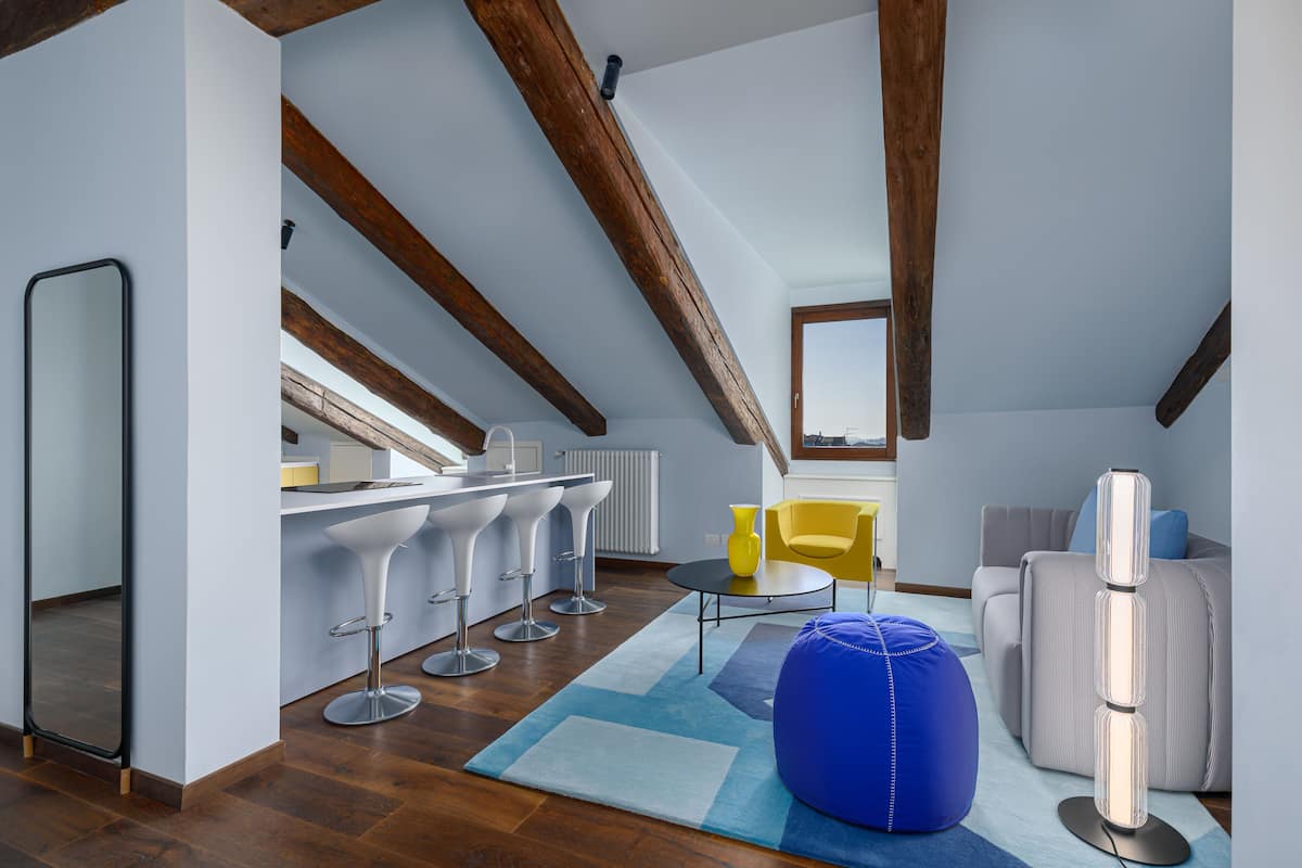 Un loft moderno con vigas de madera expuestas, una cocina con taburetes de bar, una mesa de centro redonda, un sillón de ante azul y un sofá gris.
