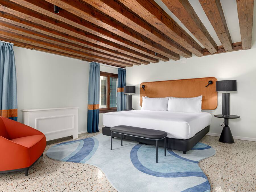 Una habitación de hotel moderna con techo de madera, una cama grande, un sillón rojo y una alfombra azul estampada.
