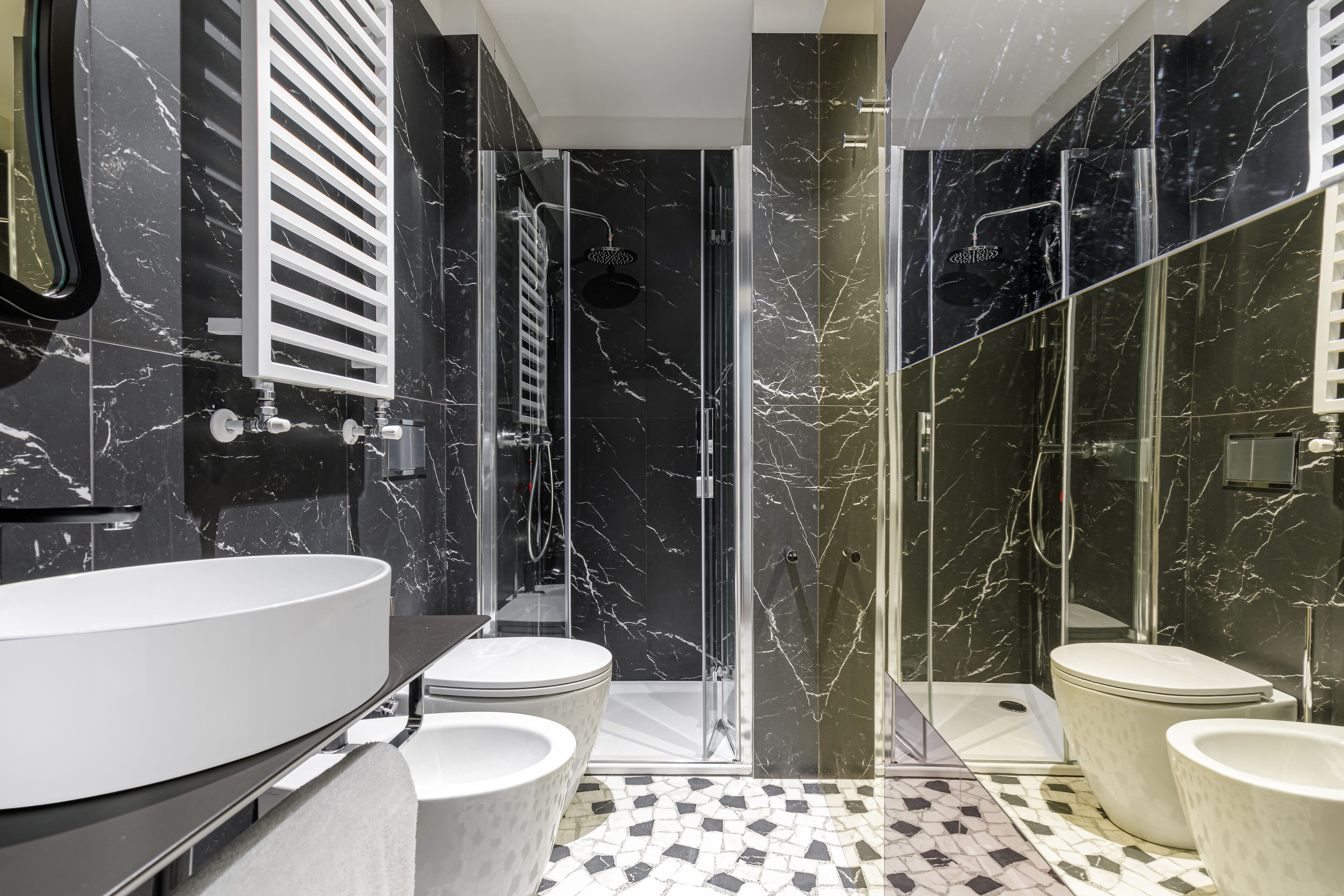 A modern bathroom with a large white sink, two toilets, a glass-enclosed shower, and a black marble wall.