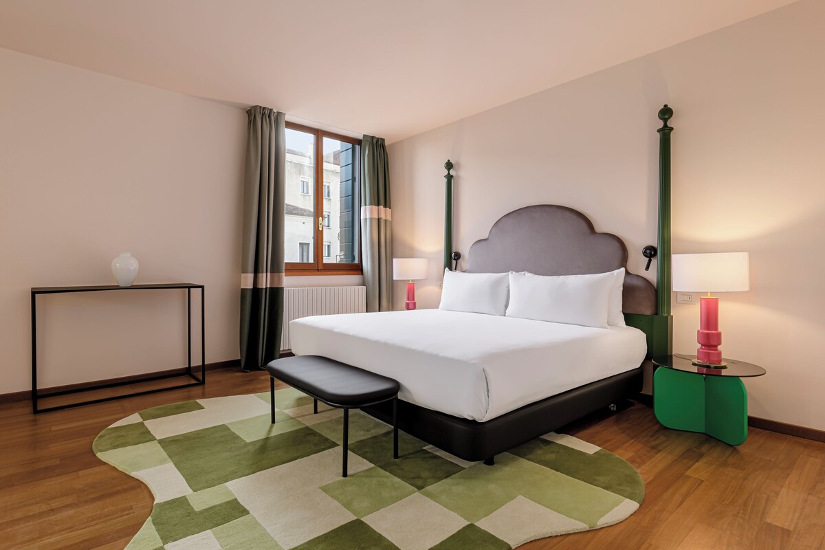 Habitación de hotel ordenada con una cama grande, mesitas de noche, un banco y vistas a la ciudad a través de la ventana.