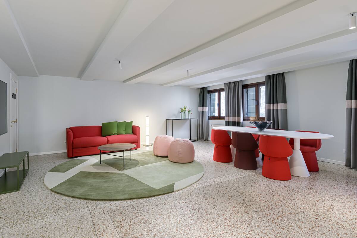 Una sala de estar moderna con un sofá rojo, otomanas verdes y rosadas, una mesa de centro redonda y un comedor con una mesa blanca y sillas rojas.