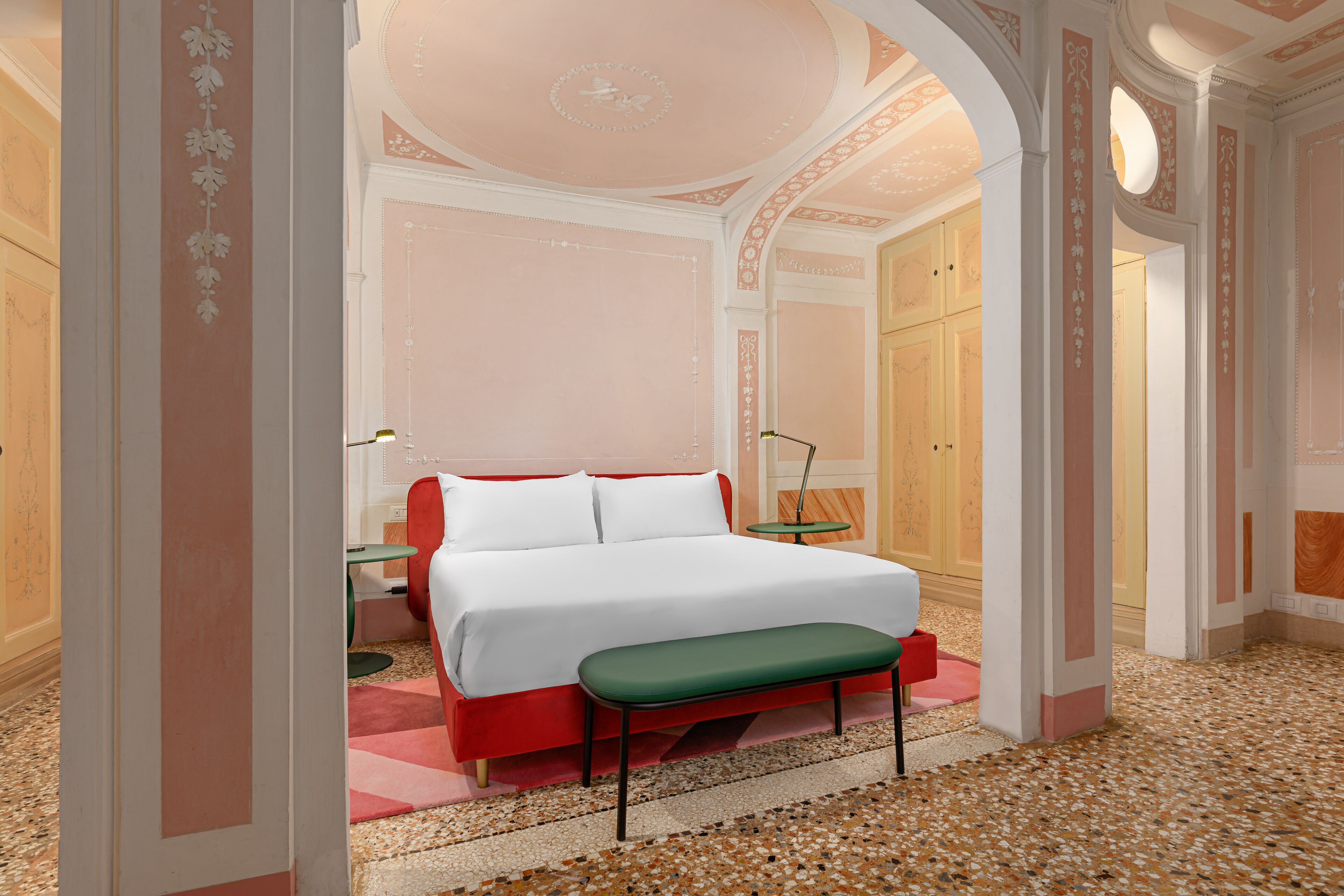 A hotel room with a large bed, a green bench, a small table, and a mirror.