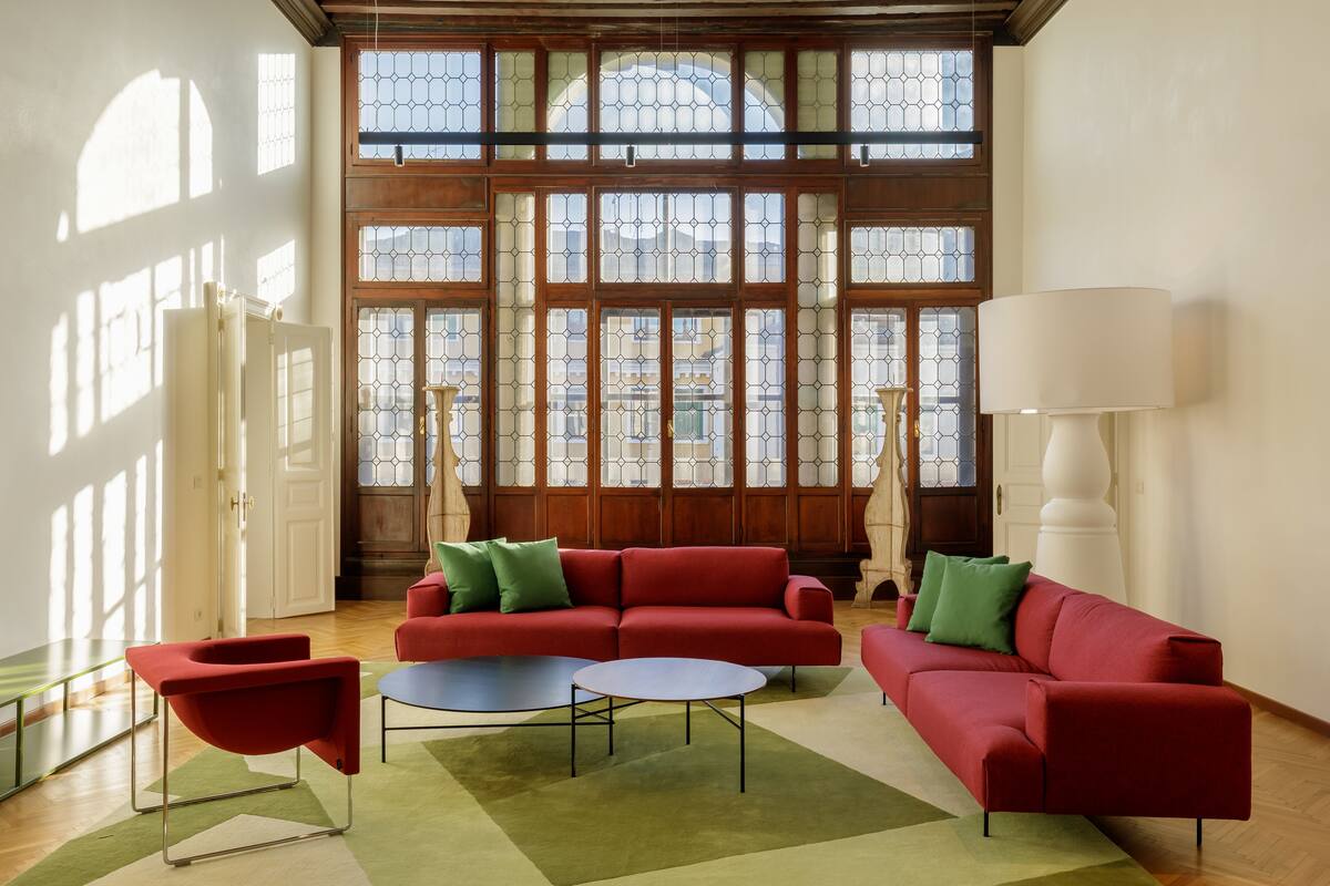 Amplia habitación con un sofá rojo, una mesa de centro redonda y una lámpara de pie. Ventanas grandes con marcos de madera y paneles de vidrio dejan entrar abundante luz natural.