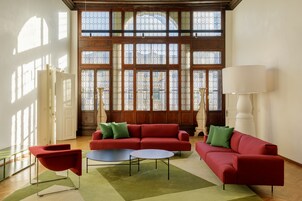 Amplia habitación con un sofá rojo, una mesa de centro redonda y una lámpara de pie. Ventanas grandes con marcos de madera y paneles de vidrio dejan entrar abundante luz natural.