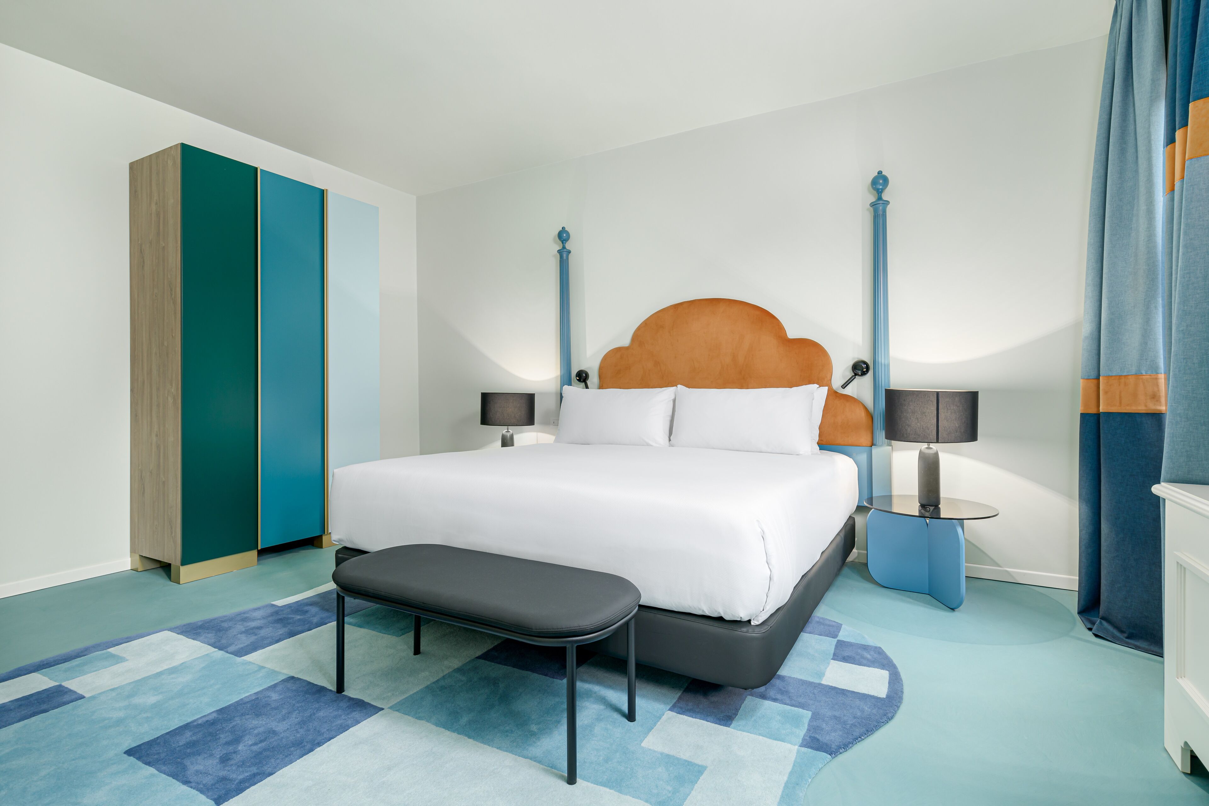 Una habitación de hotel moderna con una cama grande, un banco, dos mesitas de noche y un armario.