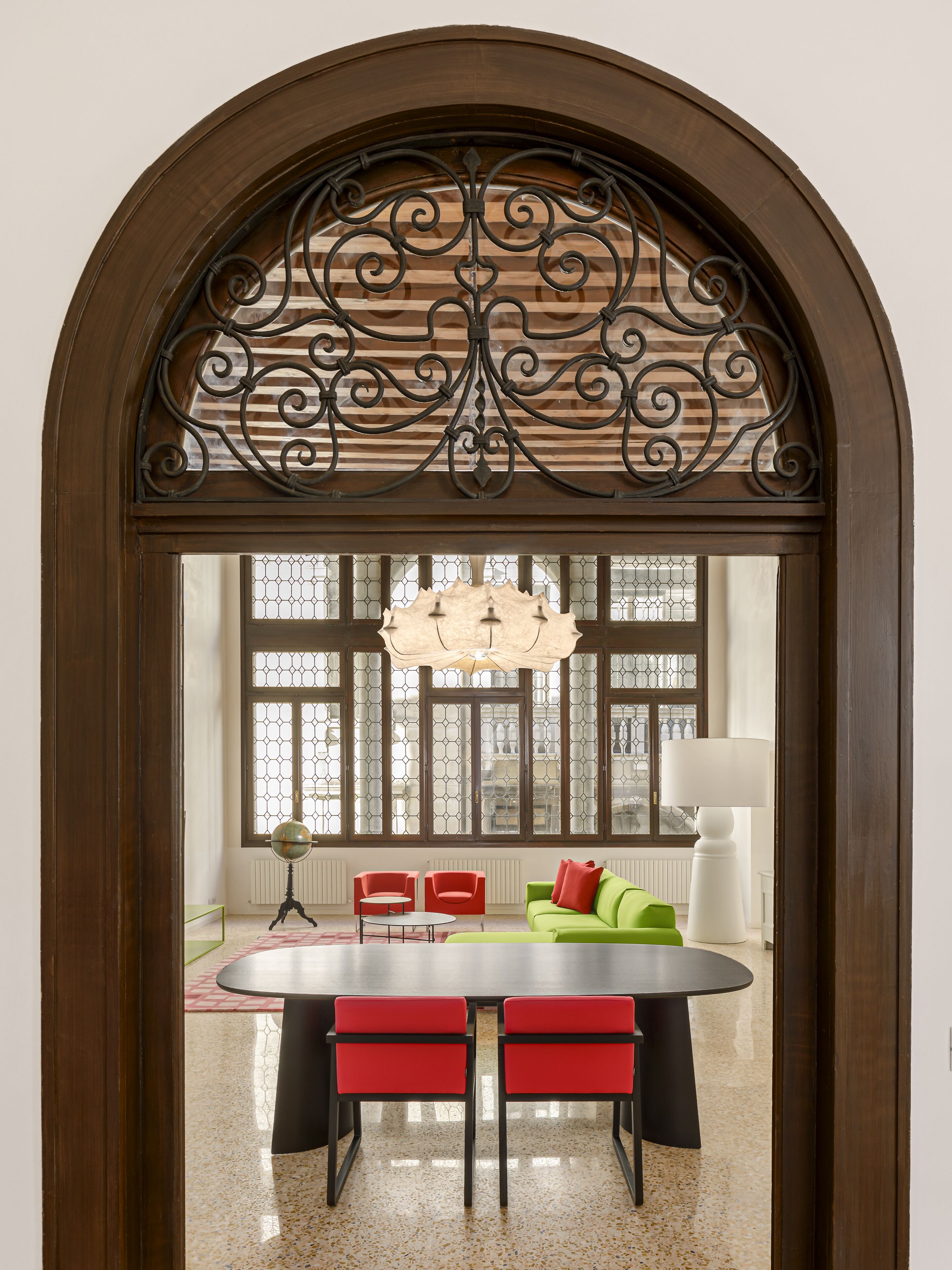 A dining area with a wooden arch, a round table, red chairs, a green sofa, and a globe on a stand.