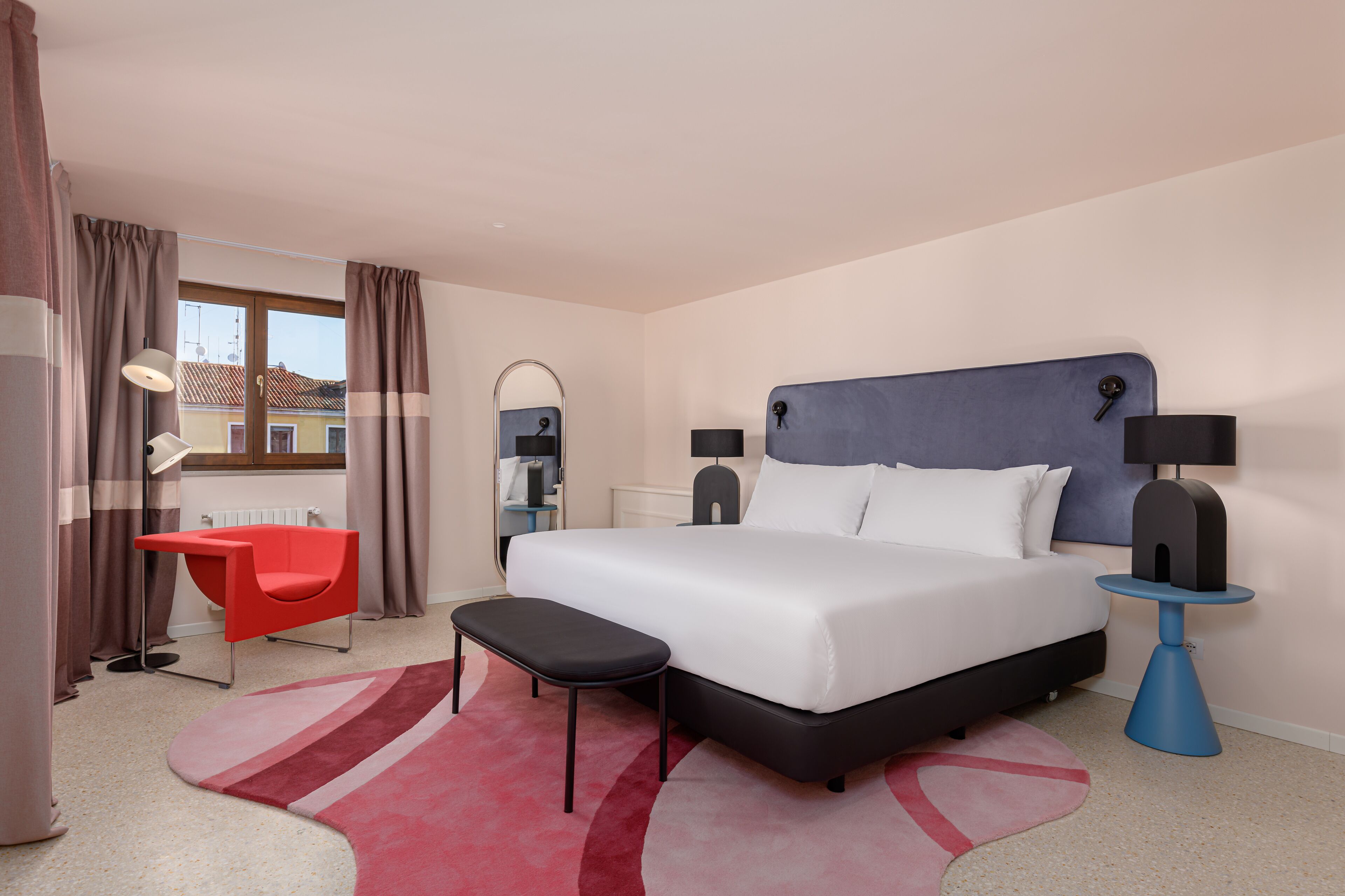 A modern hotel room with a large bed, a red chair, a small table, and a view of the outside through a window.