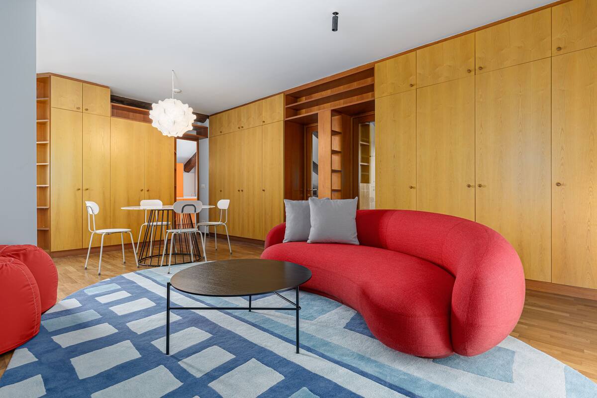 Una sala moderna con un sofá rojo, una mesa de centro redonda y un tapete azul estampado. Hay una zona de comedor con una mesa y sillas blancas, y armarios de madera al fondo.