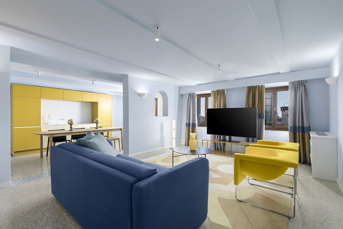 Una sala de estar moderna con un sofá azul, sillas amarillas, un televisor de pantalla plana y una cocina al fondo.