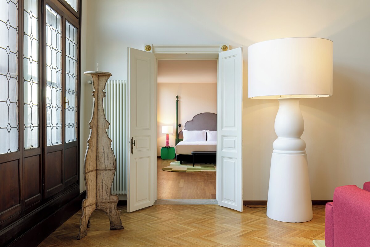 Une pièce avec un plancher en bois, une lampe blanche, un lit avec une tête de lit grise et une chaise rouge.