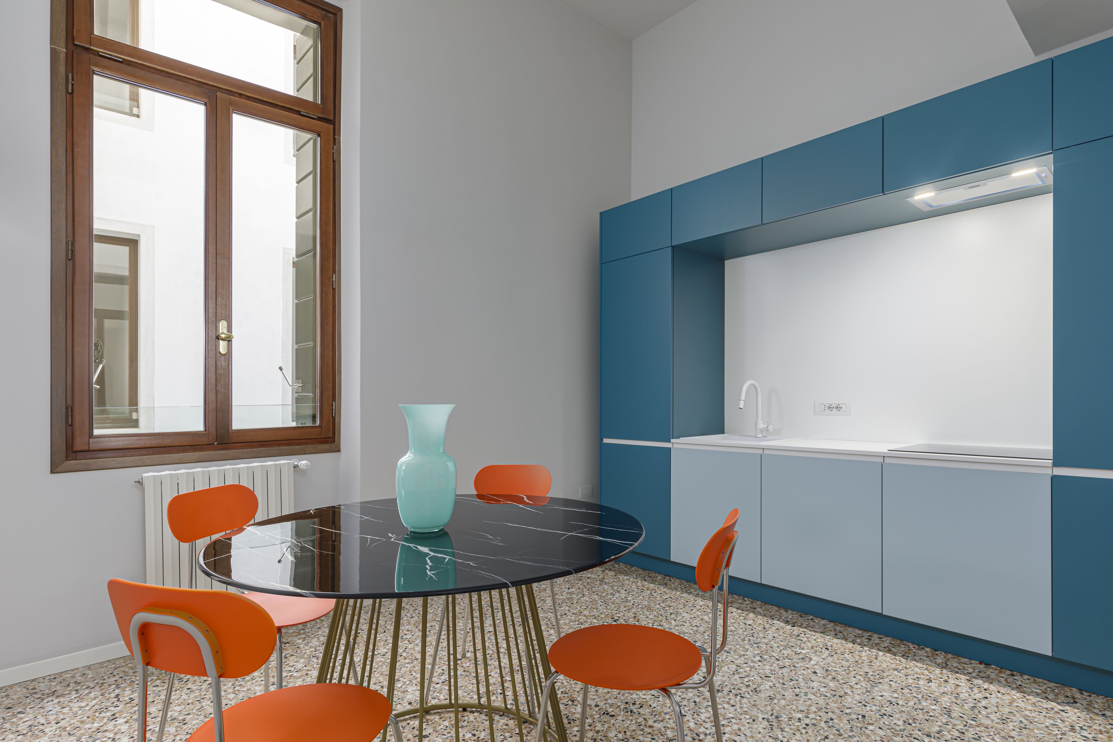 A modern kitchen with a round table and orange chairs, a blue cabinet, and a window with a view.