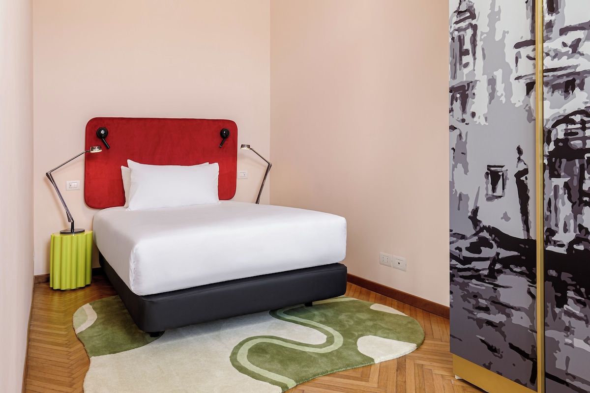 A neatly made bed with a red headboard, a green side table, and a patterned rug on a wooden floor.