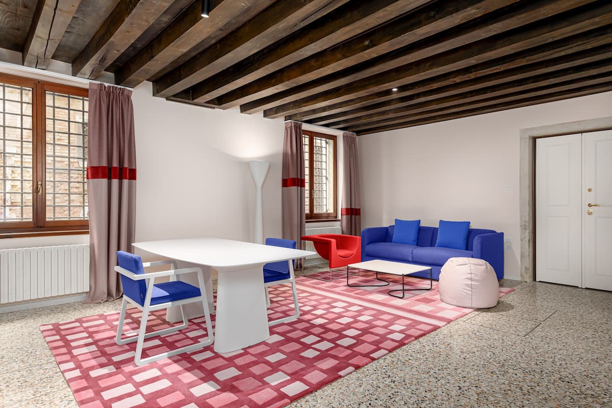 Una habitación con una mesa blanca, sillas azules, una silla roja, un sofá azul y una puerta blanca, además de una alfombra con estampado rojo y blanco.