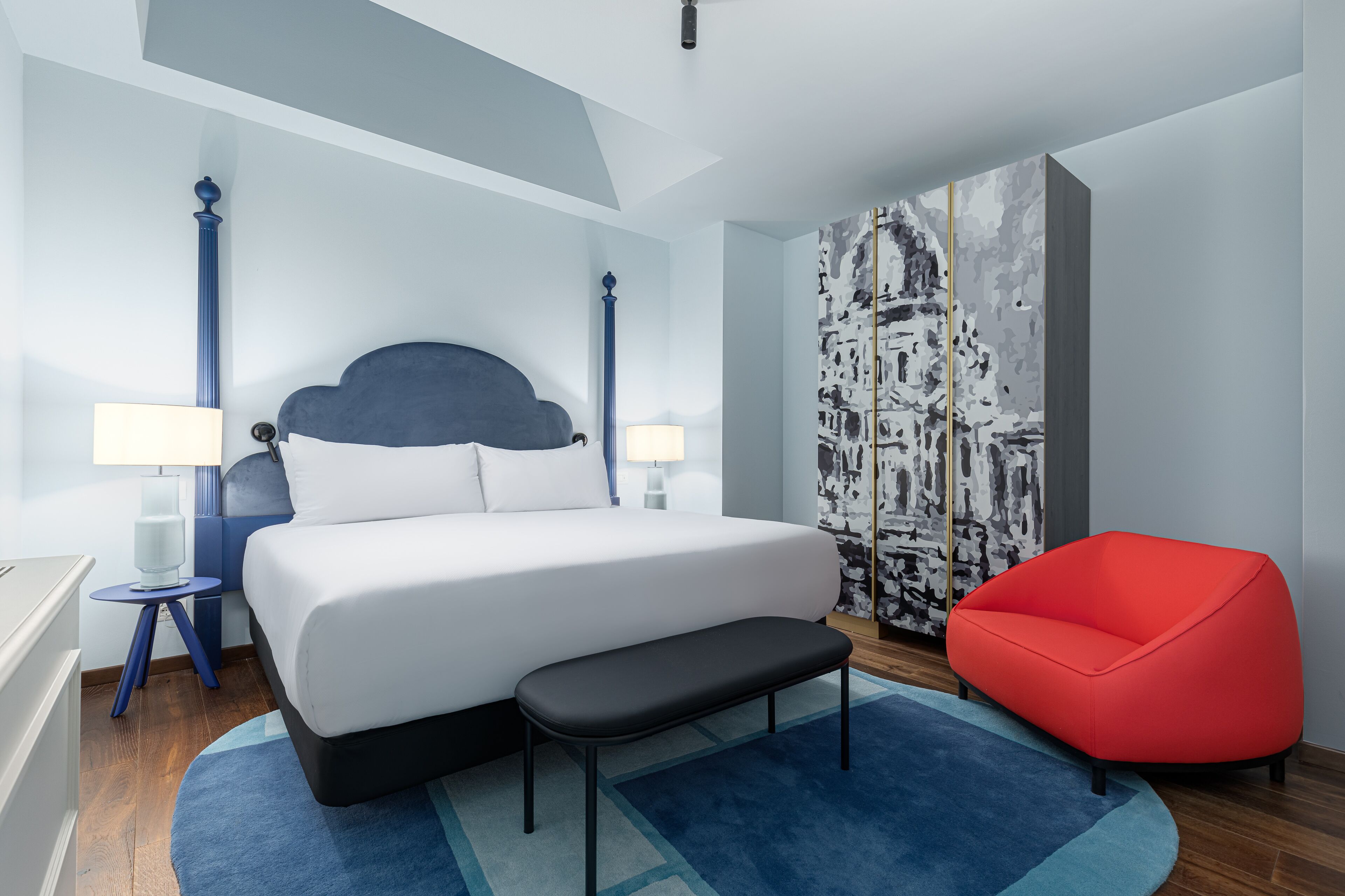 A modern hotel room with a large bed, a red armchair, and a large abstract painting.