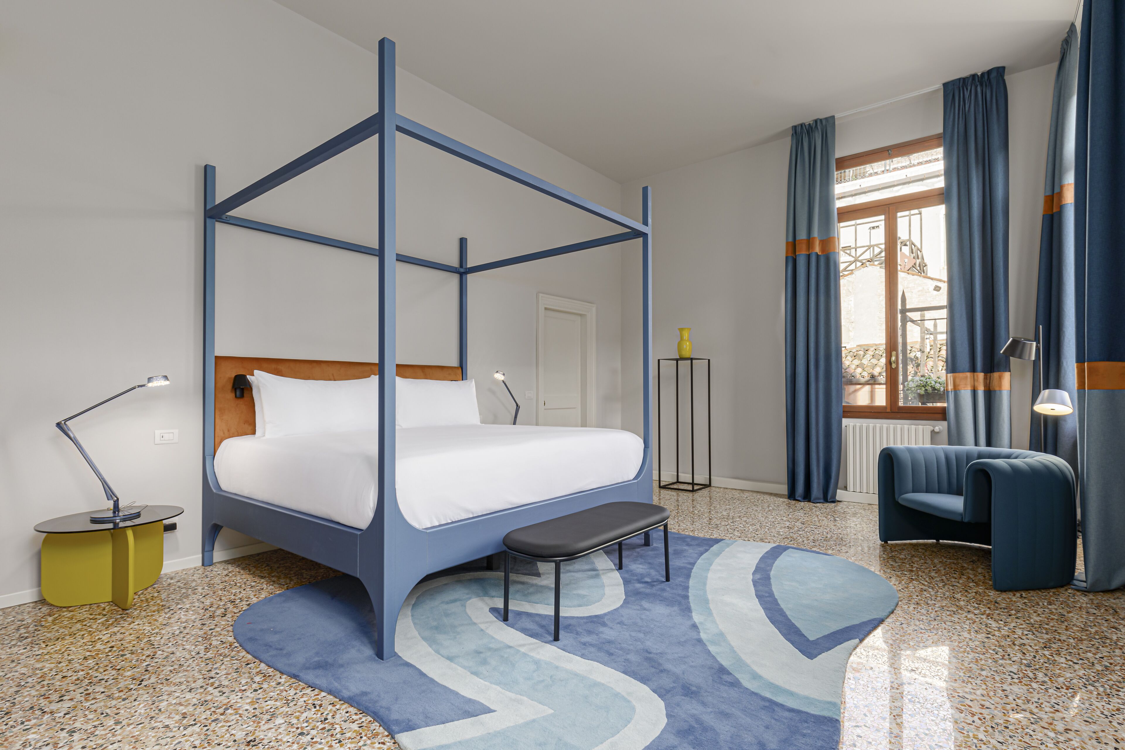 A modern hotel room with a four-poster bed, a bench, a desk with a lamp, a chair, and a view of the outdoors through a window with curtains.