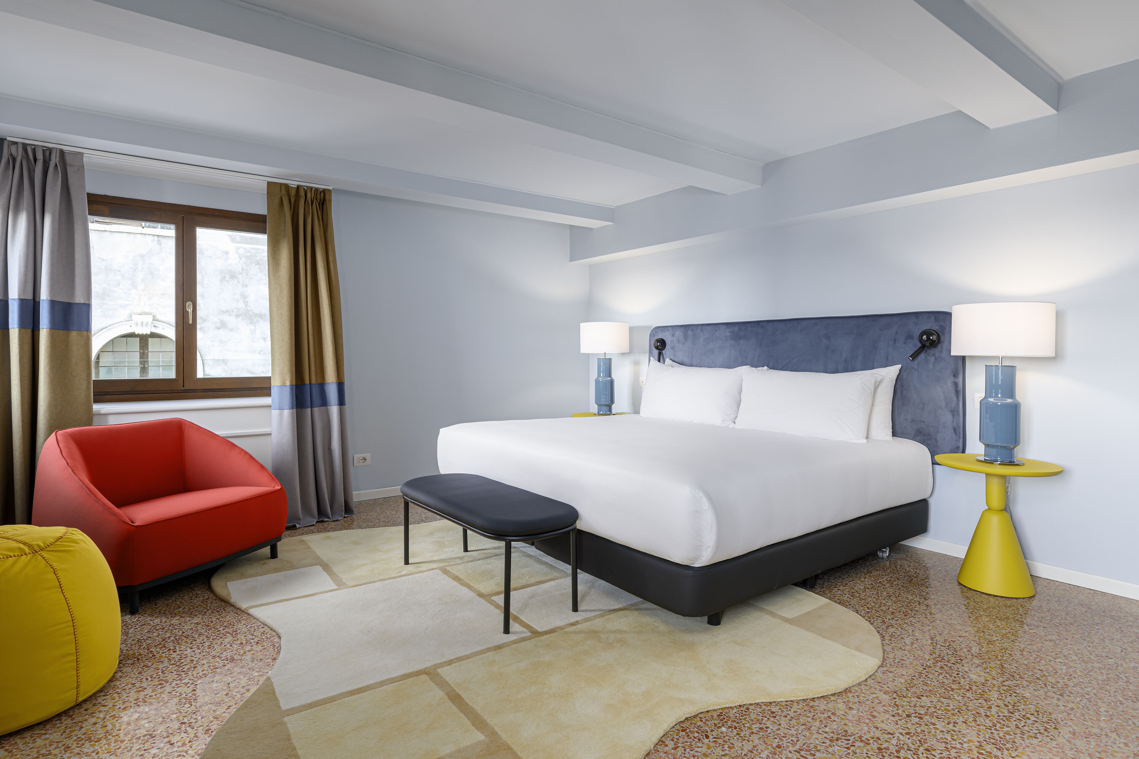Una camera d'hotel con un grande letto, una poltrona rossa, un poggiapiedi giallo e un comodino con lampade.