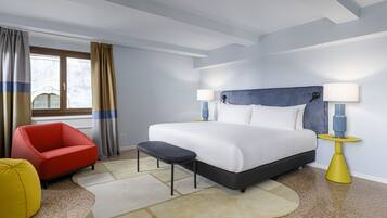 Una camera d'hotel con un grande letto, una poltrona rossa, un poggiapiedi giallo e un comodino con lampade.