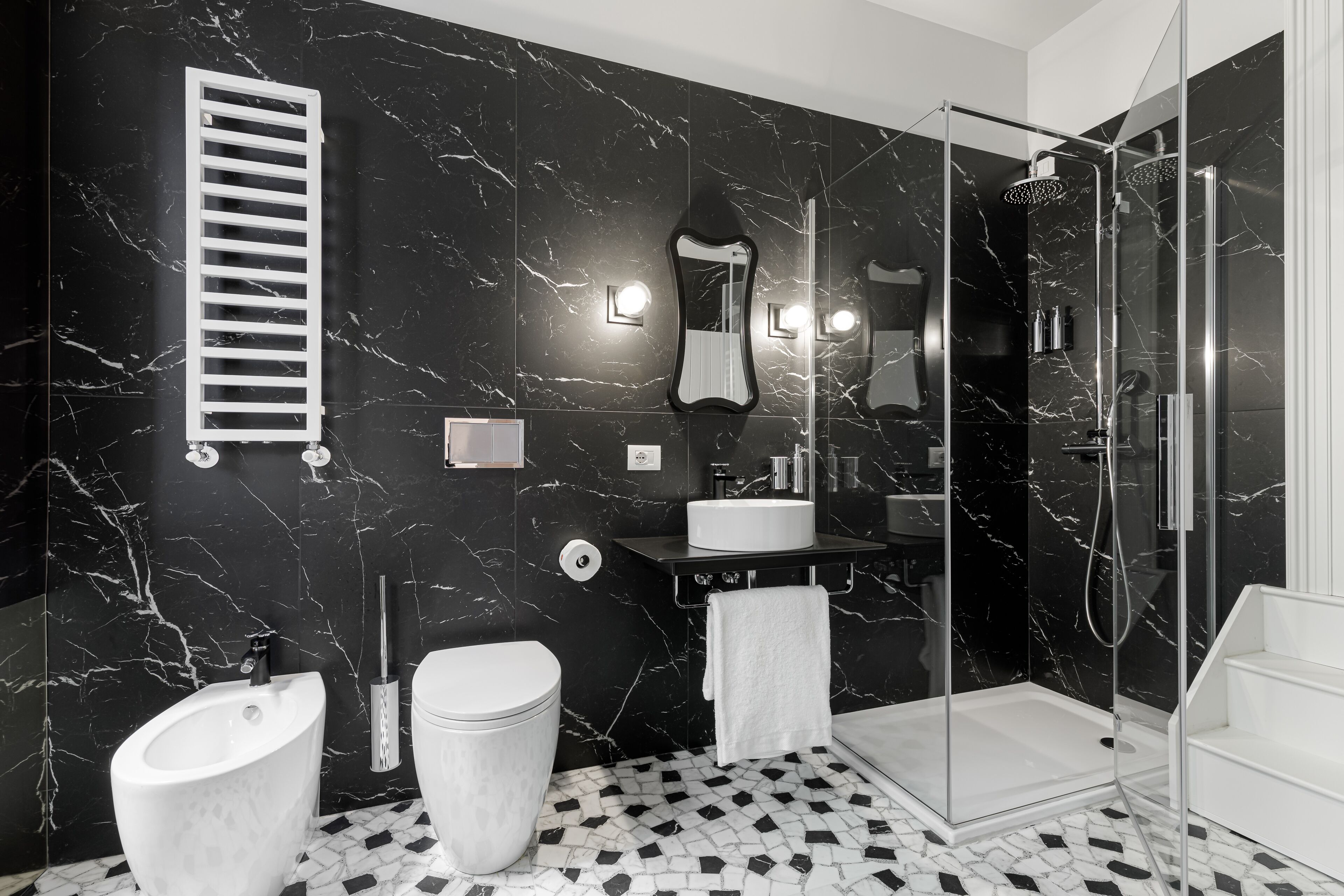 Um banheiro moderno com parede de mármore preto, box de vidro, pia branca e um bidê.