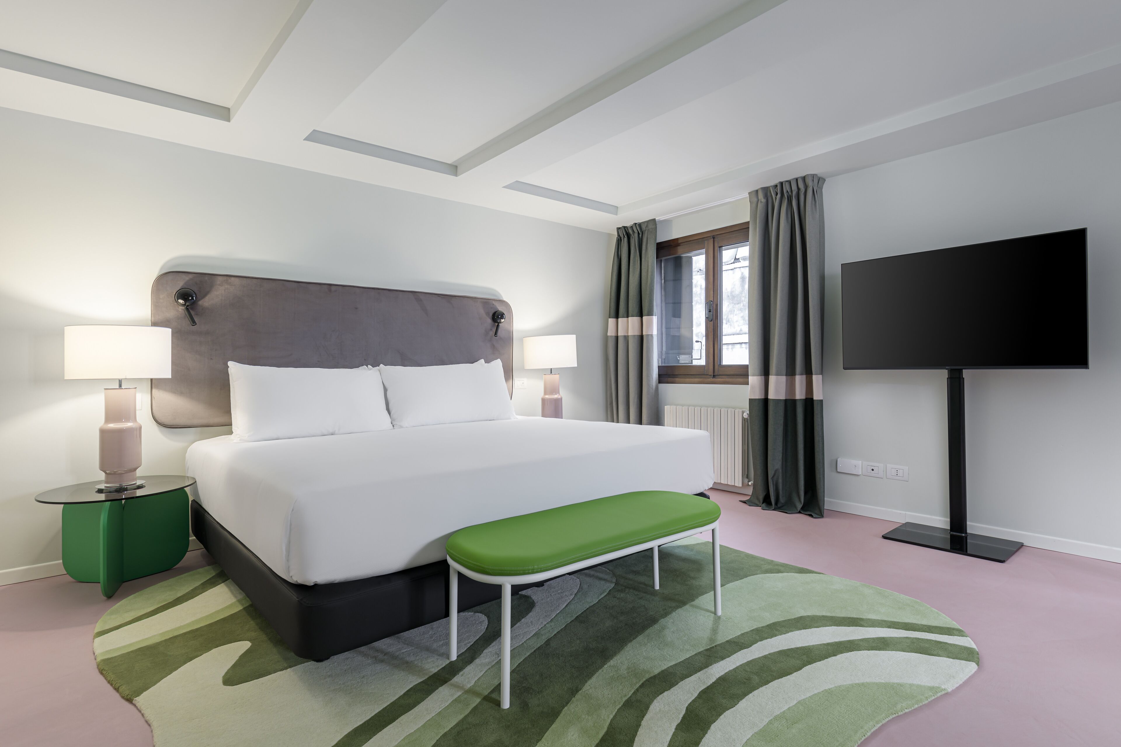A modern hotel room with a large bed, bedside lamps, a bench, a flat-screen TV, and a view of the outdoors.
