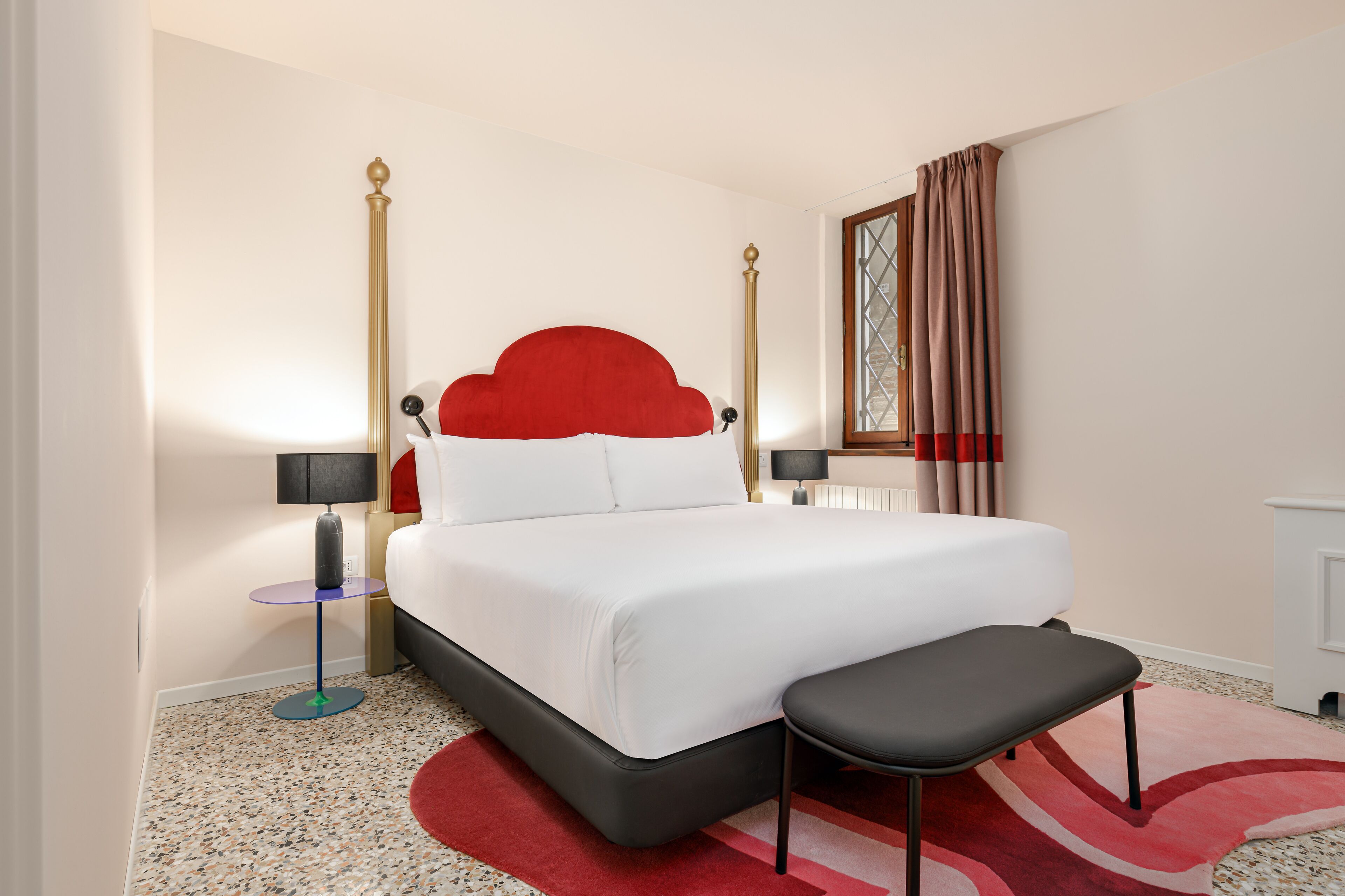 Habitación de hotel ordenada con una cama grande, una mesita de noche pequeña, un banco y una ventana con cortinas.
