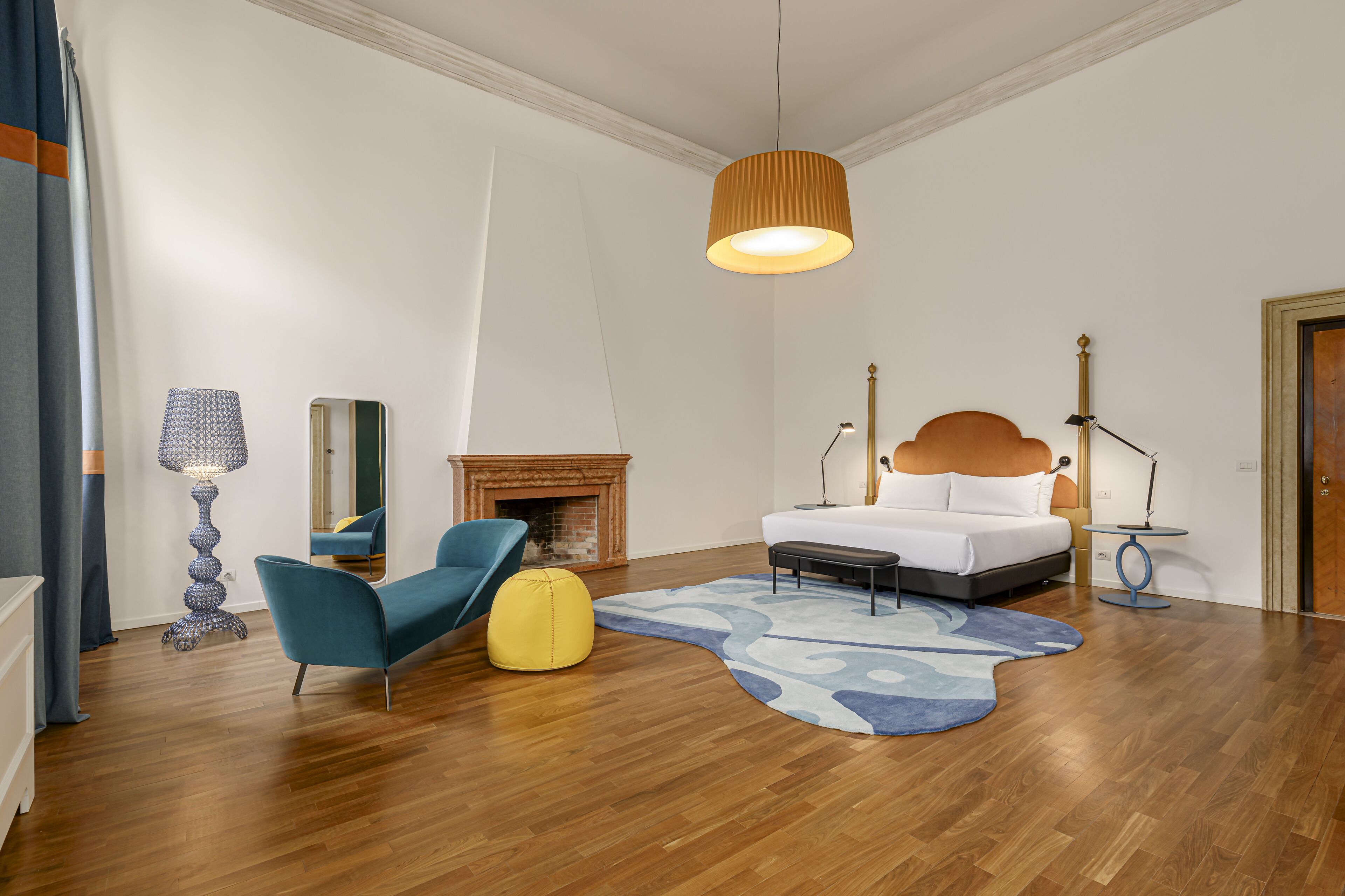 A modern bedroom with a bed, a blue chair, a yellow ottoman, a fireplace, and a hanging light fixture.
