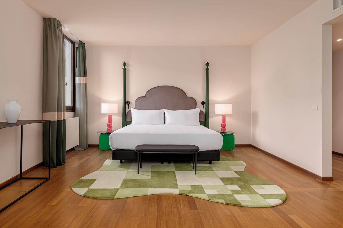 Habitación de hotel ordenada con una cama grande, lámparas de noche, un banco, alfombra verde y una ventana con cortinas.