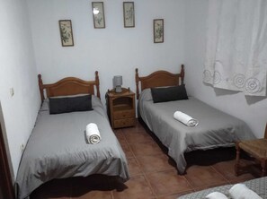 Une chambre avec deux lits en bois, chacun recouvert d’une couverture grise et agrémenté d’une serviette enroulée. Une table de chevet avec une lampe se trouve entre les deux lits. Les murs sont unis et ornés de trois tableaux encadrés.