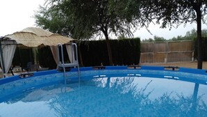 Una piscina exterior con interior azul, rodeada de árboles y una zona cubierta con techo de paja.