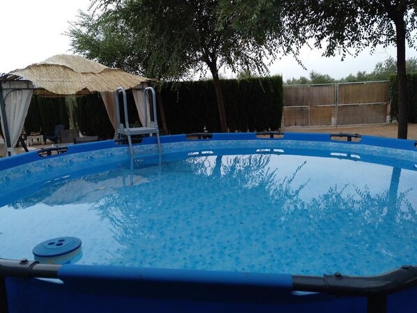 Une piscine extérieure aux parois bleues, entourée d’arbres et dotée d’un espace abrité avec un toit de chaume.