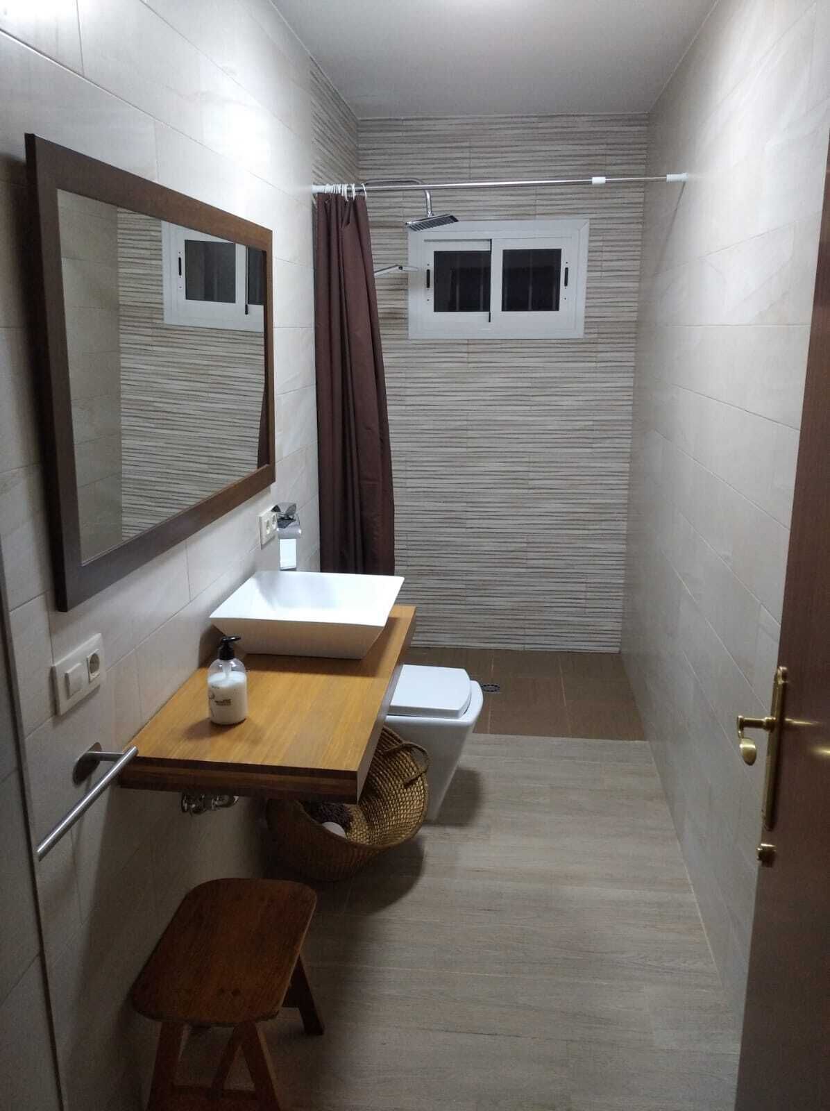 Une petite salle de bain avec un meuble-lavabo en bois, un miroir, un lavabo blanc, un espace douche et un petit tabouret.