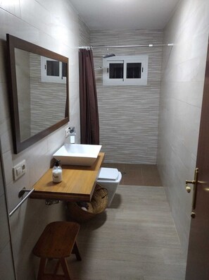 Une petite salle de bain avec un meuble-lavabo en bois, un miroir, un lavabo blanc, un espace douche et un petit tabouret.