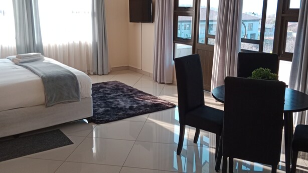 Una habitación de hotel con cama, televisión, mesa de comedor con sillas y ventanas grandes con cortinas.