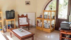 A living room with a wooden chair, a table with a lace tablecloth, a bookshelf, a radiator, a television, and a window with a view of trees. - Group Accommodation in Crete Separate Houses (Apokoronas)