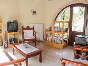A living room with a wooden chair, a table with a lace tablecloth, a bookshelf, a radiator, a television, and a window with a view of trees. - Group Accommodation in Crete Separate Houses (Apokoronas)