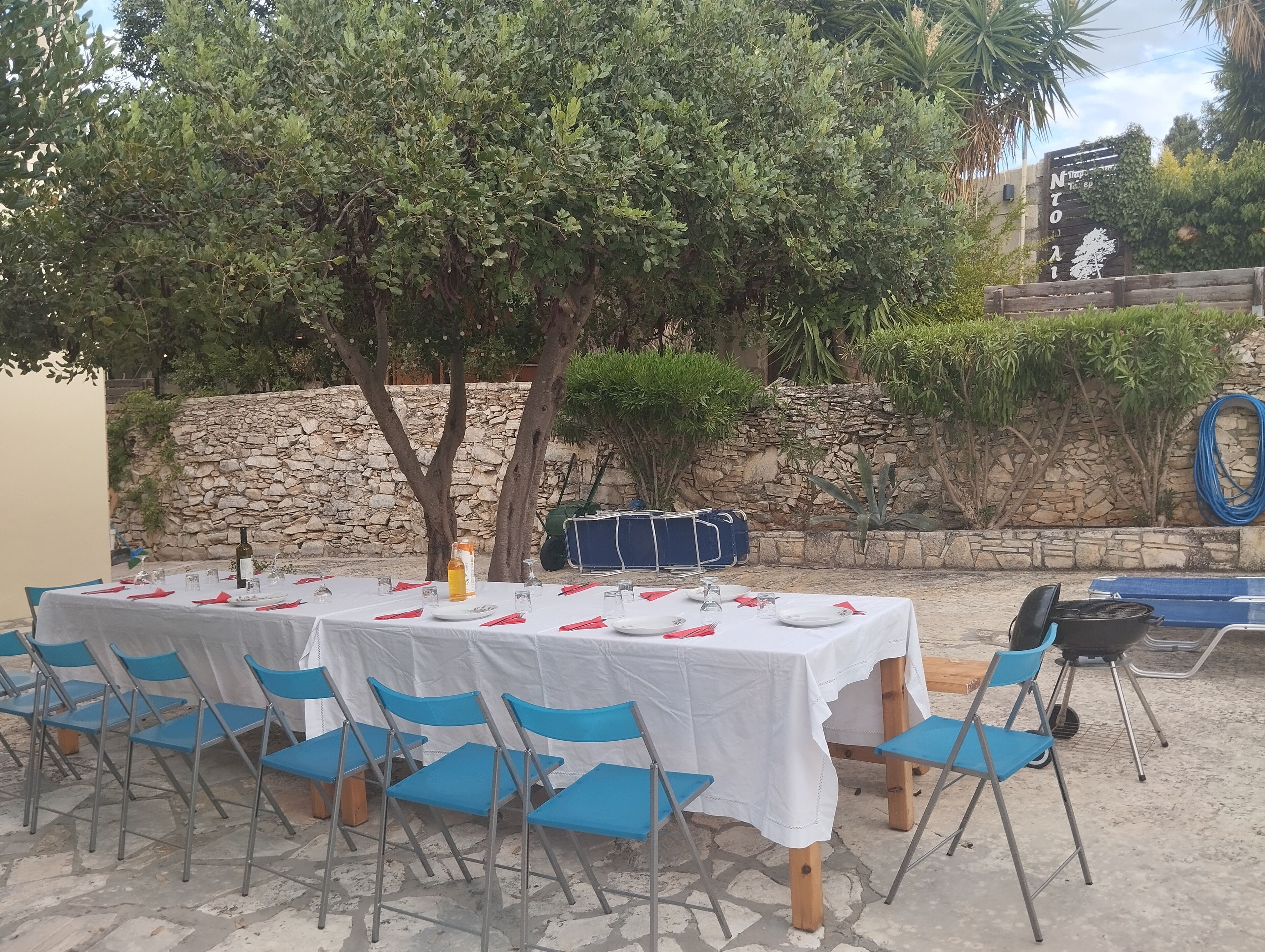 Un espacio de comedor al aire libre con mesas preparadas para cuatro personas, sillas azules y una pared de piedra de fondo.
