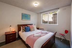 A neatly made bed with a pink blanket, a wooden nightstand with a lamp, a red heart-shaped chair, and a window with blinds. - Beautiful 2-bed Unit in St Kilda West w/ Parking (St Kilda West)