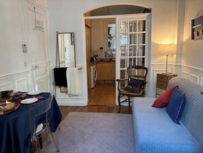 A room with a bed, a chair, a table, and a mirror. There is a door leading to another room with a washing machine.