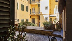 Un patio con edificios amarillos, un banco de metal, plantas en macetas y un regador de metal.