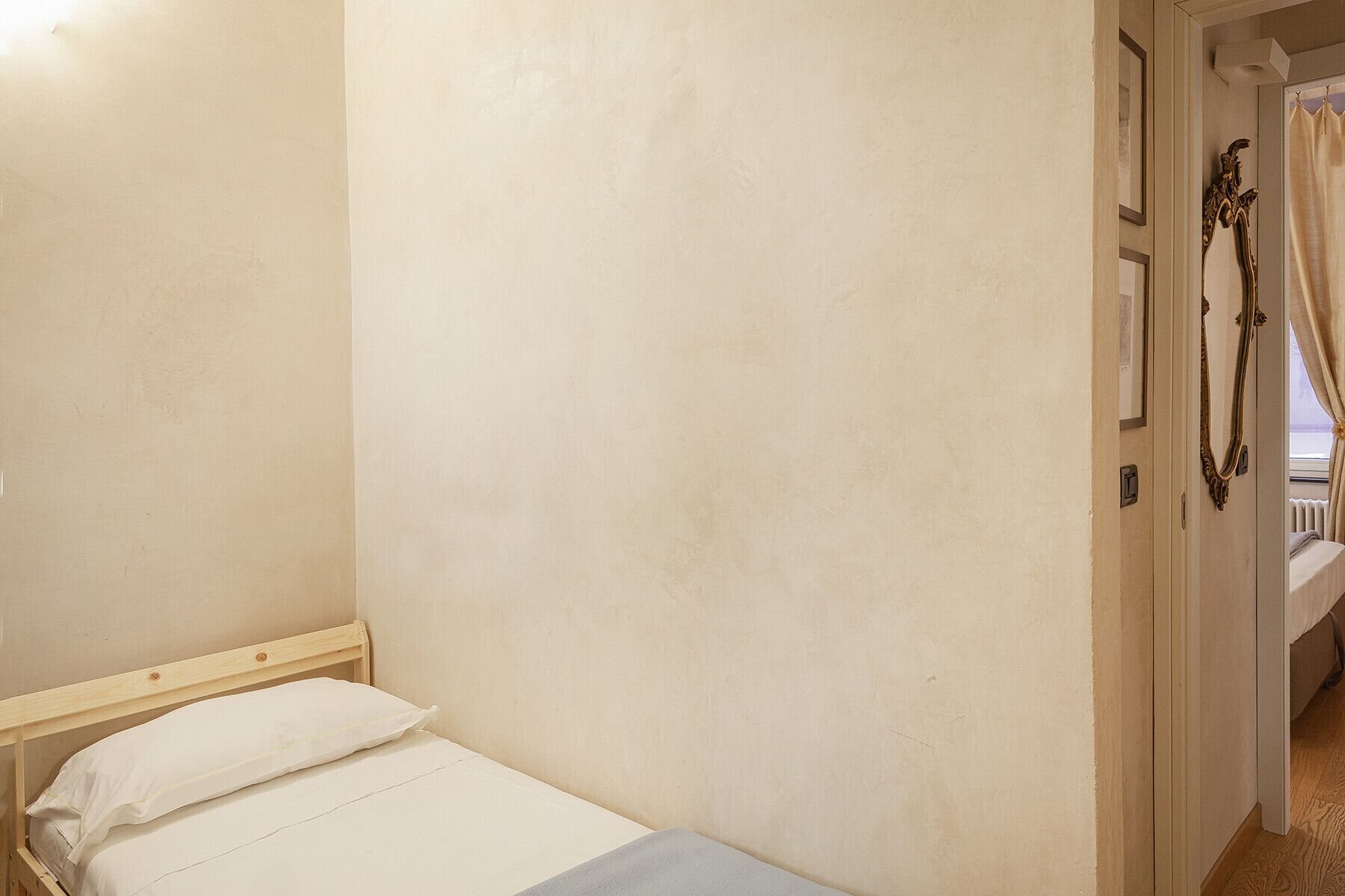 A small, single-bed room with a wooden headboard, a plain wall, and a door with a mirror and curtains.