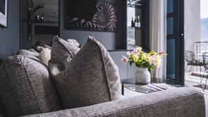 Ein modernes Wohnzimmer mit einem grauen Sofa, einem Glastisch und einer Vase mit Blumen. An der Wand hängt ein großes gerahmtes Kunstwerk, und durch eine Glastür ist ein Blick ins Freie möglich.