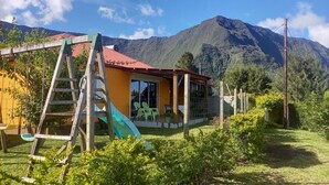 A house with a red roof, a wooden ladder, and a green slide in a garden with mountains in the background.