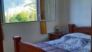 A bedroom with a wooden bed, a bedside table, a nightstand, a window with a view of greenery, and a ceiling fan.