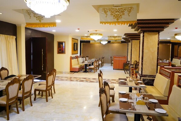 A hotel dining area with multiple tables and chairs, chandeliers, and a reception desk.
