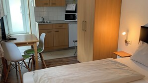Habitación de hotel pequeña con cama, escritorio con silla, kitchenette y armario.