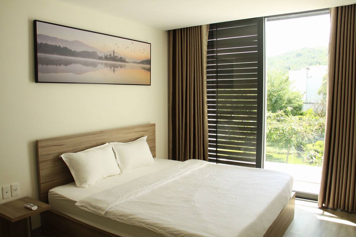 A hotel room with a large bed, a nightstand, a remote control, and a window with blinds.