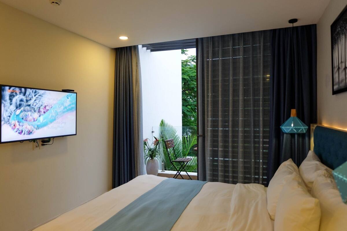 A modern hotel room with a large bed, a flat-screen TV mounted on the wall, a balcony with a view of greenery, and a decorative lamp.