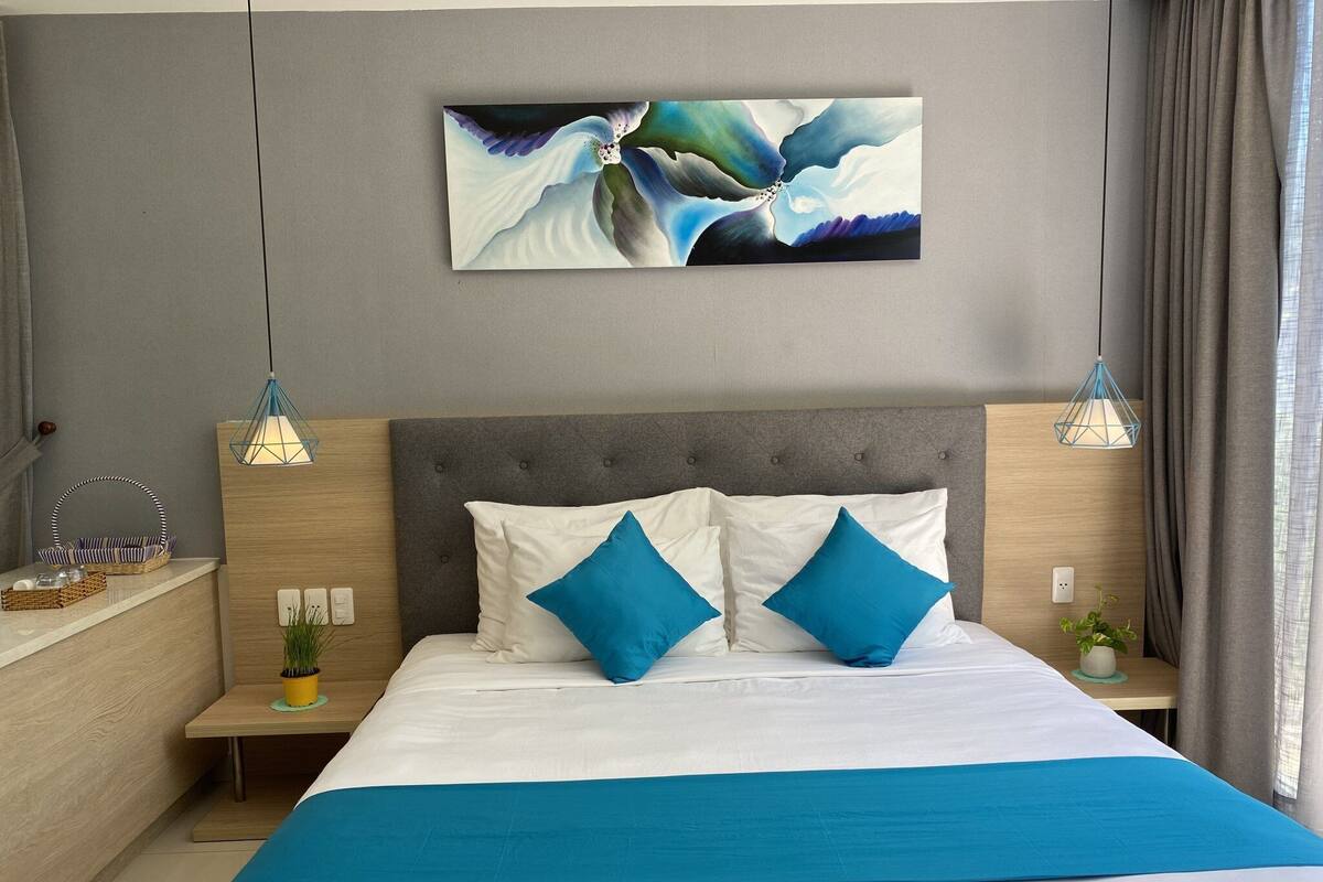 A modern bedroom with a grey upholstered headboard, a large bed with white and blue bedding, two bedside tables with lamps, and a wall-mounted abstract painting.