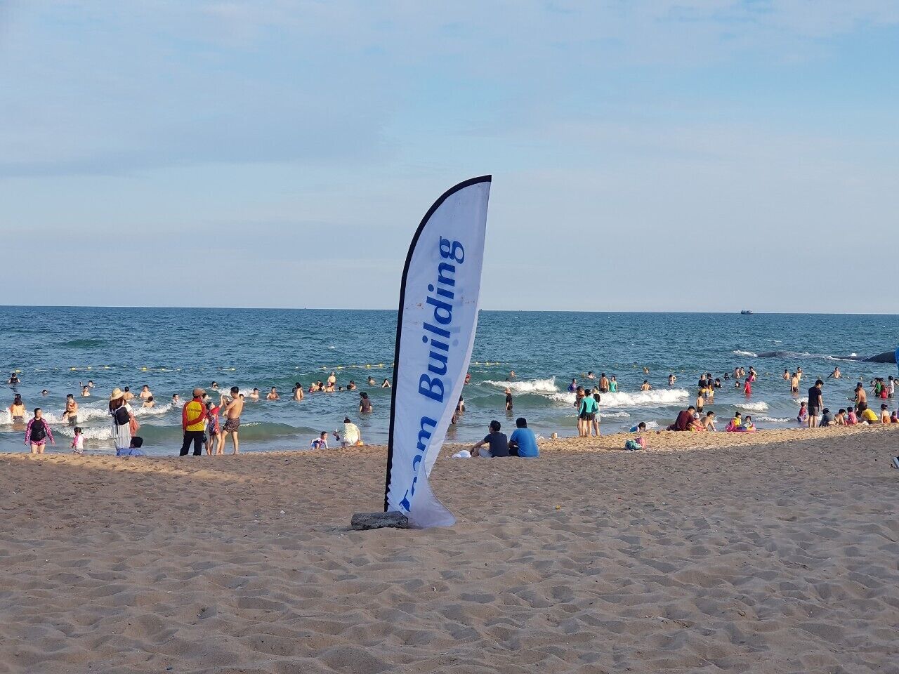 A beach with a flag reading "Team Building" and people swimming and relaxing on the sand.