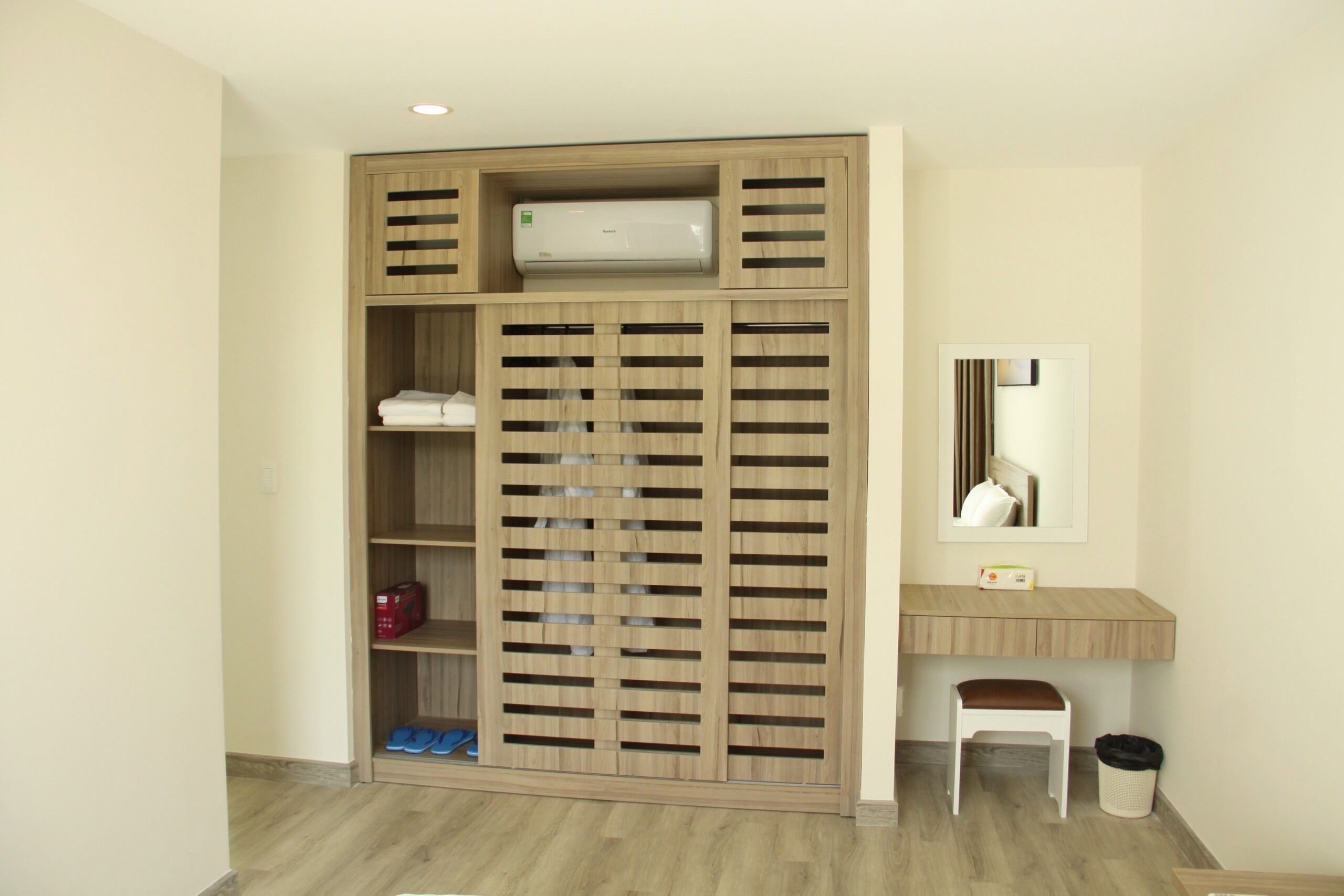 A room with a wooden wardrobe, a small desk, a mirror, and a trash bin.