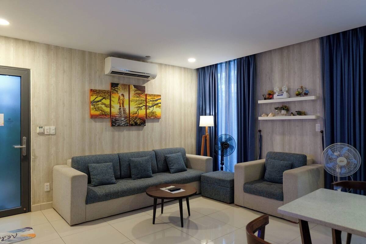 A modern living room with a beige sofa, a matching armchair, a small table, and a fan. There are two wall-mounted air conditioners, a painting, and a window with blue curtains.